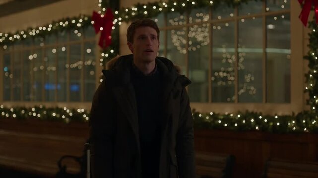 Christmas travel man waiting outside decorated station at night with suitcase wearing warm winter parka jacket