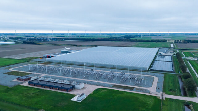 Aerial view of 150kv-station middenmeer adjacent to a large greenhouse complex with wind turbines in the background in the flat landscape of Middenmeer, Noord-Holland, Netherlands.