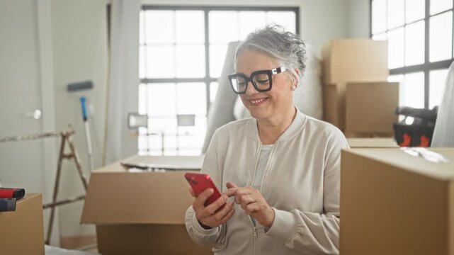 Woman with short hair and glasses tapping a red smartphone with hands while seated amid cardboard moving boxes and packing supplies in building; contentment transition.