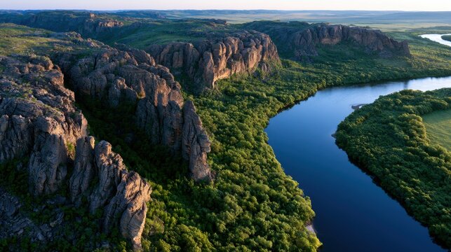 Aerial view of a winding river flowing through verdant forest and rugged rocky cliffs under a clear sky in a vast natural landscape