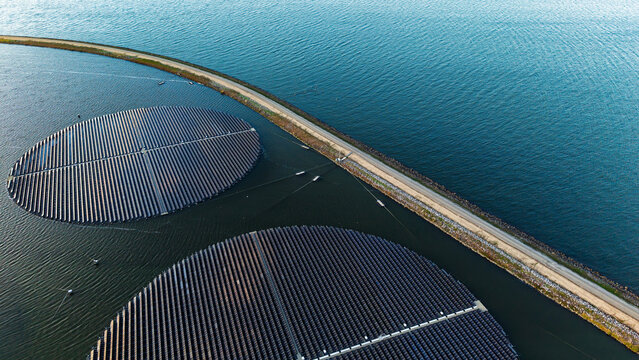 Aerial view of circular floating solar panels arranged in large arrays on a calm lake beside a long embankment in the Netherlands.