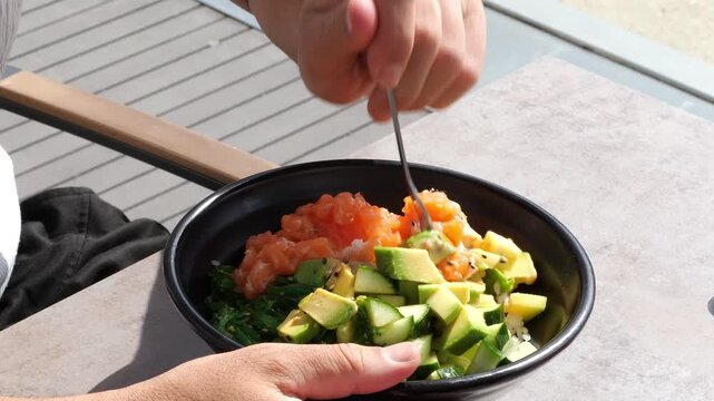 Hands Eating Fresh Hawaiian Salmon Poke Bowl with Fork Diced Avocado Cucumber and Rice at Outdoor Table Healthy Lunch Food Lifestyle Concept