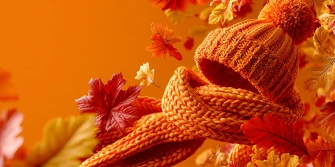 Cozy orange knitted beanie and scarf surrounded by vibrant autumn leaves against a warm orange background, evoking fall comfort and seasonal warmth. © Anastasiya