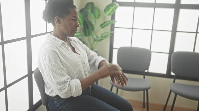 Woman seated on chair with hands resting on knees, waiting in a clinic building by a grid window and spare chairs, subtle pensive posture; concern.