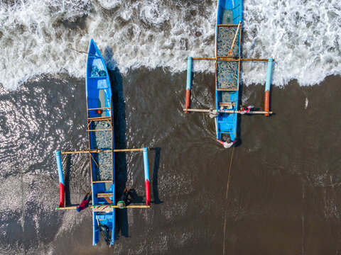 Aerial view of two traditional blue outrigger fishing boats resting on the dark sandy shore with fishermen working nearby as waves wash onto the beach in Cilacap, Central Java, Indonesia.