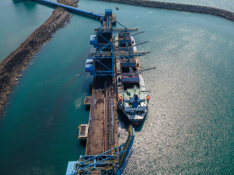 Aerial view of a large cargo ship docked at a port terminal with industrial loading cranes, surrounded by turquoise water and a breakwater in Cilacap, Central Java, Indonesia.