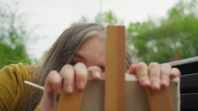 Close up shows person working on art project outdoors. Hands grip frame of small canvas set up in front of them while they hold and move paintbrushes focusing intently on their work