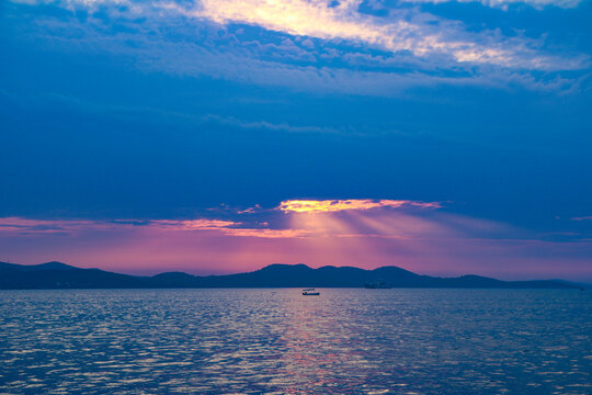 View of a small boat on the calm Adriatic Sea at sunset with sunbeams breaking through clouds over the hills of Hvar, Split-Dalmatia County, Croatia.
