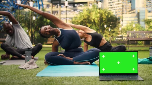 Isolated green screen appears on laptop with replaceable display during yoga class as participants do side stretch exercise close to device, supporting screen replacement and yoga in urban park.