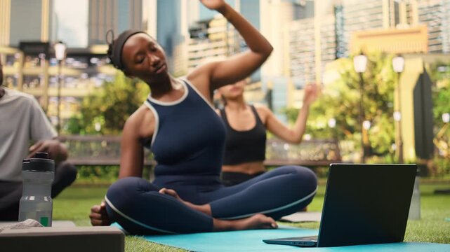Young woman does a side stretch exercise in front of laptop with friends, following a digital yoga activity session with participants in city park while stretching together outdoors.