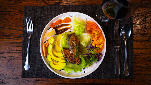 View of a plate of grilled fish with avocado, lettuce, corn, and carrots served on a wooden table in Indonesia.