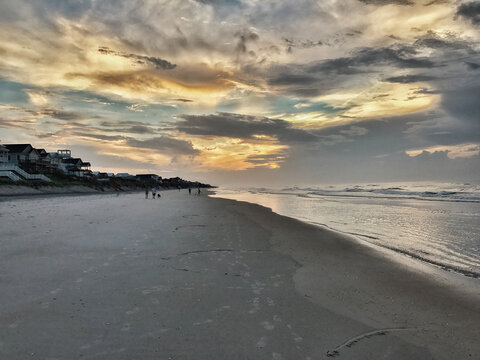 View of the sandy shoreline of Topsail Island at sunset with dramatic clouds, calm ocean waves, and coastal houses perched on the dunes in North Topsail Beach, North Carolina, United States.