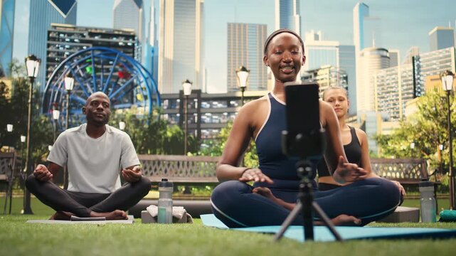 Yoga instructor extends arms while group follows on mats during streaming, filming live wellness practice with participants in urban park.