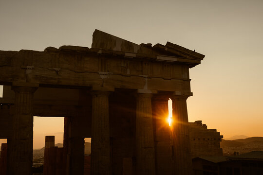 View of the Acropolis in Athens at sunset, with the sun shining through the ancient stone columns of the temple, casting a warm golden glow over the historic ruins in Athens, Greece.