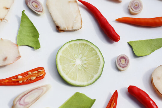 A colorful, close-up pattern of Thai herbs: lime slices, galangal, lemongrass, chilies, and kaffir lime leaves.
