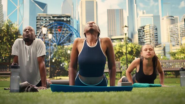 Woman instructor holds cobra pose on exercise mats with participants during synchronized outdoor yoga class, stretching back muscles against urban public park, supporting flexibility and wellness.