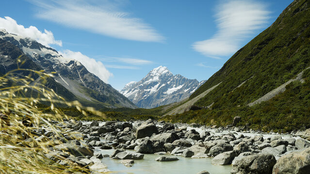 View of Hooker Valley track with the snow-capped Aoraki Mount Cook peak, rocky riverbed, and green slopes under a blue sky in Mt Cook National Park, Canterbury Region, New Zealand.