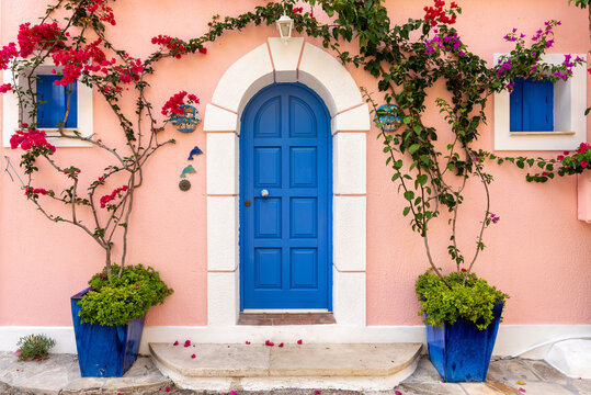 View of a vibrant blue arched door set in a pastel pink wall, framed by lush bougainvillea flowers and blue shutters in Asos, Greece.