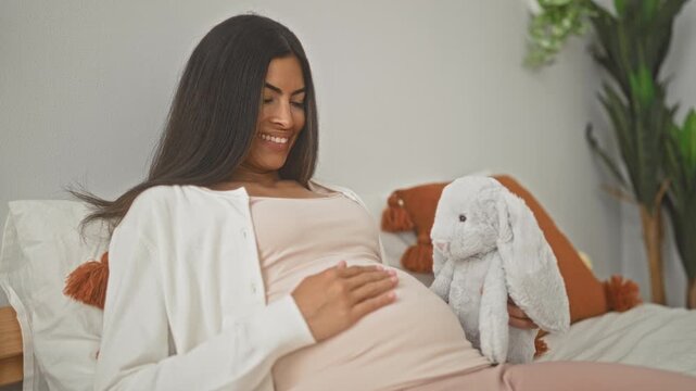 Woman smiling, touching her pregnant belly while holding a soft plush bunny on a bed in a building; maternal serenity.