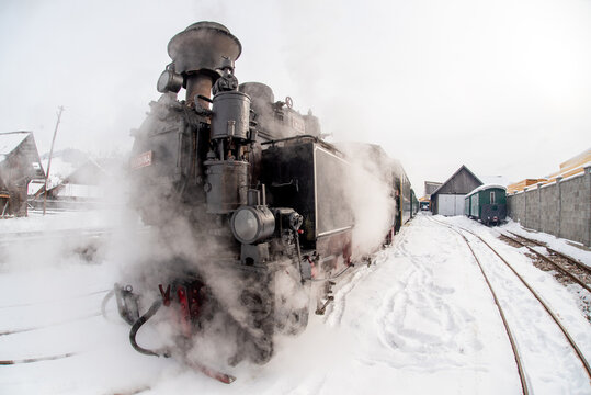 View of the Mocanita steam train emitting thick steam while standing on snowy tracks at the station in Moldovita, Suceava County, Romania.