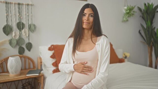 Pregnant woman cradling belly on bedroom bed with hand on bump and boho decor; serene anticipation hope.