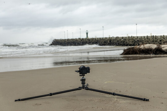 View of a camera mounted on a tripod on a sandy beach with a breakwater and crashing waves in the background in Debki, Pomeranian Voivodeship, Poland.