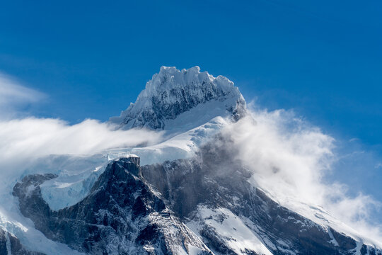 View of the snow-capped peak of Cordillera del Paine surrounded by swirling clouds against a clear blue sky in Torres del Paine, Magallanes, Chile.