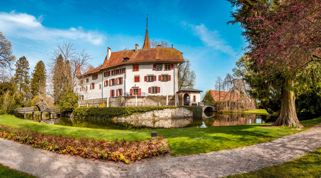 View of Schloss Landshut, a historic moated castle with white walls and red shutters, surrounded by a lush green lawn and a calm pond under a clear blue sky in Utzenstorf, Bern, Switzerland.