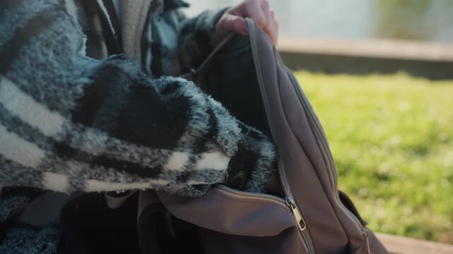 Person in checkered coat sits outdoors on grass and wood. They use their right hand to unzip main compartment of brown backpack revealing its interior lining while holding it open with both
