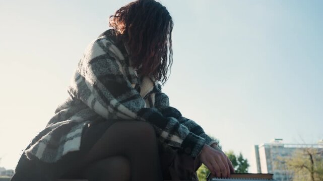 Woman sits outdoors reading book in sunlight. Woman wears casual clothing including tights has long hair with sunlit strands carries brown backpack and holds open book. scene includes trees
