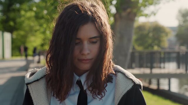 Woman with long brown hair wears white collared shirt and black tie under jacket as woman walks along path beside water. scene is set in daylight featuring green trees sunlight filtering