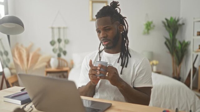 Man typing on laptop at wooden desk with hands on keyboard and holding mug in a studio setting with bed and plants visible; focused productivity.