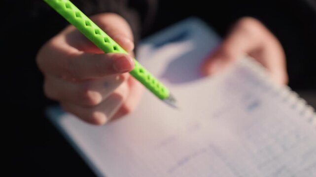 Close up view shows person hands gripping bright green mechanical pencil above open spiral bound notebook with faint blue grid lines. focus remains on texture of skin pen details and paper