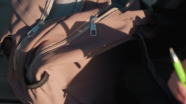 Close up view of pink fabric backpack featuring zippers handle details and texture. hand holds green pen near bag in soft lighting with shadows zipper