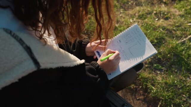 Woman writes notes and solves math problems on graph paper while sitting outside. scene shows hands holding pens over open spiral bound notebook filled with handwritten text including graphs