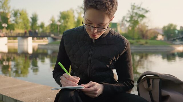 Young woman sits outdoors near water writing notes on paper. scene includes calm greenery in background with soft daylight and natural composition notebook