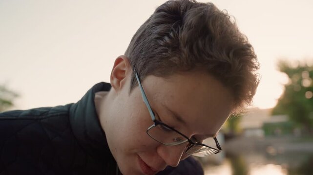 Man with short hair wears black framed glasses while looking away from camera. Man is dressed in dark quilted jacket standing outdoors near water and trees at sunset. scene features soft