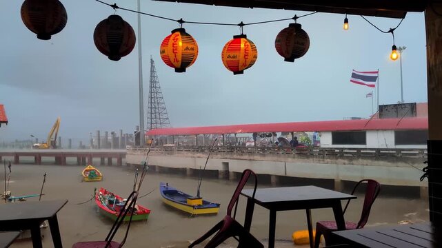 Ko Lan harbour seen from waterfront restaurant interior with Chinese lanterns, string lights, colorful fishing boats, red roofed pier and Thai flag under stormy overcast sky, static shot.