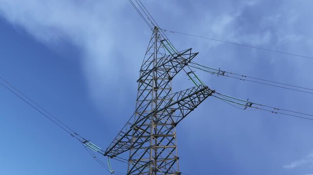 Tall steel electricity pylon carries multiple overhead transmission lines and green insulators against a bright blue sky with drifting clouds overhead