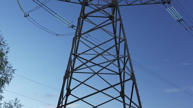 Low angle view moves upward along a steel transmission tower, revealing lattice supports, suspended insulators, power lines, and clear blue sky