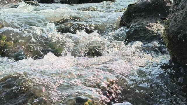 Fast-flowing river water moving between natural rocks and stones, creating dynamic ripples, small waves, and foamy patterns that highlight the movement and texture of a natural freshwater stream.