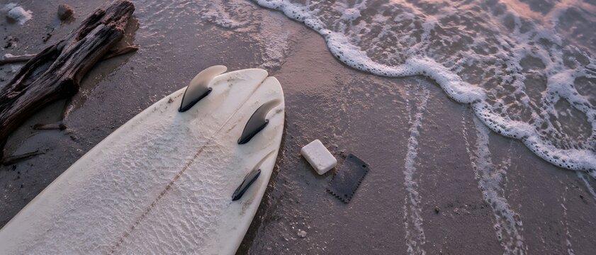 Surfboard lying fins-up on wet sand with a wax comb and a bar of paraffin beside the deck