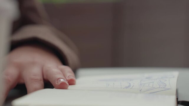 Close up shows person hand gripping blue pen and moving it across page of open notebook actively sketching or writing. scene focuses on detailed texture details like skin tone variations