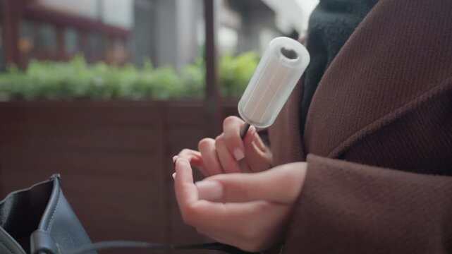 Woman hand holds electric device likely for personal care or grooming. background is softly blurred suggesting outdoor urban setting with greenery and building elements. Hands are steady