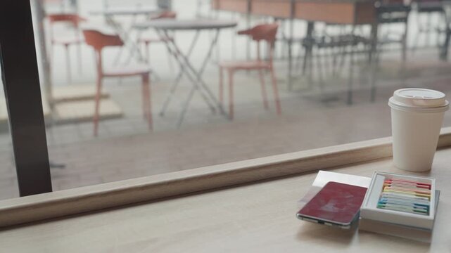 Woman draws at table by large windows. Woman uses watercolors to sketch details into small notebook with art supplies and coffee nearby. scene is bright indoors near empty cafe