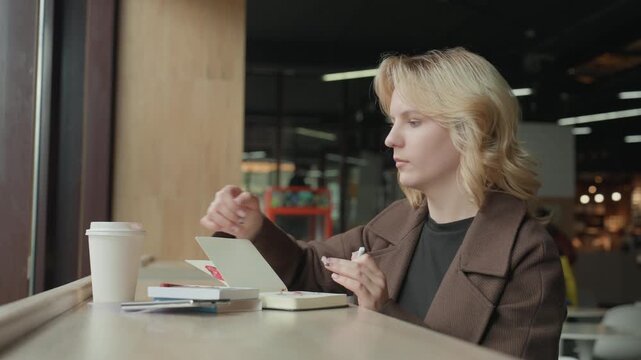 Woman sits at wooden table in what appears to be cafe or casual indoor setting. Woman interacts with objects on table including opening envelope and holding white paper cup