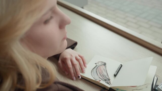 Young lady with blonde hair draws bird sketches on paper while resting her head near table. Woman wears brown jacket and black top focusing intently as woman adds details to existing drawing