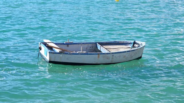 An empty wooden rowboat with an oar rocks gently on rippling turquoise water in a sheltered bay, moored beside a small yellow buoy. A peaceful maritime scene for summer travel and coastal leisure.