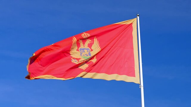 A red and gold Montenegro flag with a crowned double headed eagle emblem flutters in the wind on a white pole against a bright blue sky, a symbol of national pride.