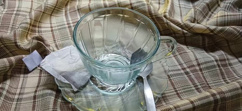 hand places two tea bags next to a clear glass cup against a background of warm, brown checkered fabric